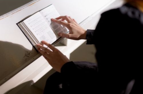 Photo of a hymnal resting on a surface, with someone's hands holding it there and seemingly following along with their right index finger.